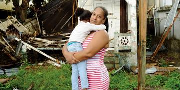 A mother holds her daughter in her arms while standing in front of destruction caused by Hurricane Maria in Puerto Rico during the 2017 hurricane. A mother holds her daughter in her arms while standing in front of destruction caused by Hurricane Maria in Puerto Rico during the 2017 hurricane.