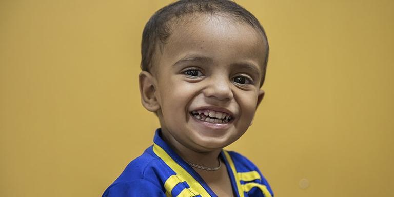 A child in a blue shirt against a yellow background smiles at the camera. A child in a blue shirt against a yellow background smiles at the camera.