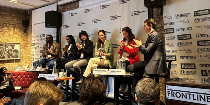 Six people sit on a panel at a speaking event with an audience Six people sit on a panel at a speaking event with an audience