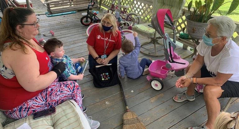 A Save the Children worker along with two other women, sit on a porch with two young kids, surrounded by toys. A Save the Children worker along with two other women, sit on a porch with two young kids, surrounded by toys.