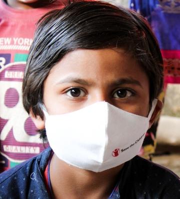 Young boy in a blue shirt looks into the camera wearing a Save the Children face mask. Young boy in a blue shirt looks into the camera wearing a Save the Children face mask.