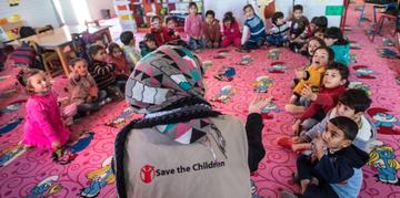 A woman wearing a Save the Children branded shirt sits with a group of children in a circle. A woman wearing a Save the Children branded shirt sits with a group of children in a circle.