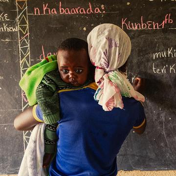 A young mother holds her son, while she writes on a blackboard at a Save the Children learning and livelihood center. A young mother holds her son, while she writes on a blackboard at a Save the Children learning and livelihood center.