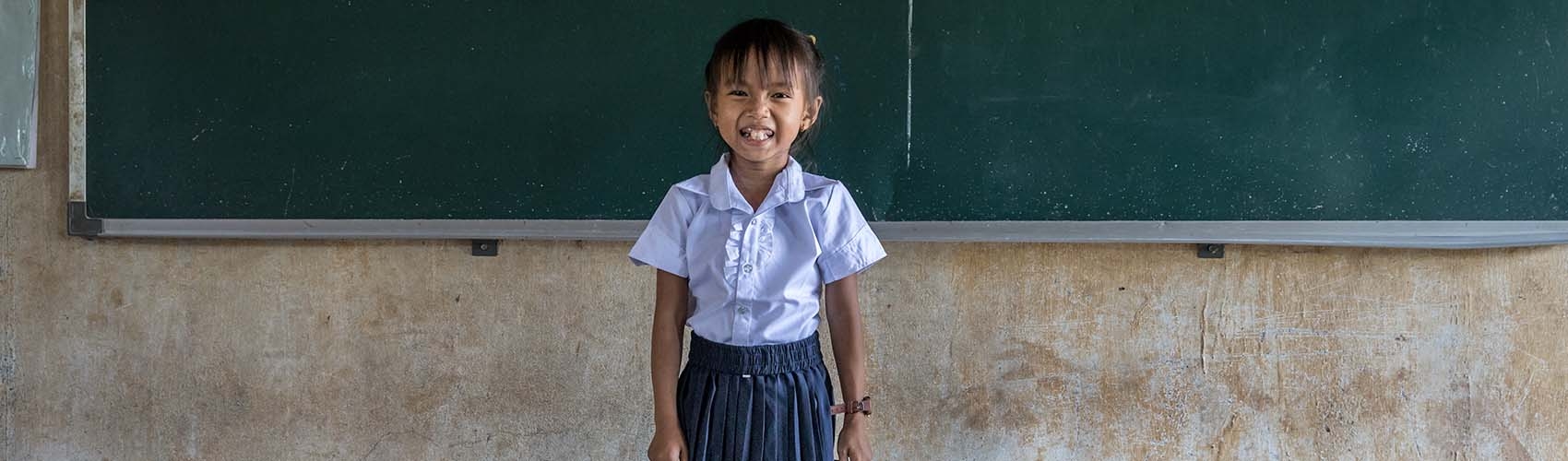 A girl in Cambodia stands in front of a chalkboard.