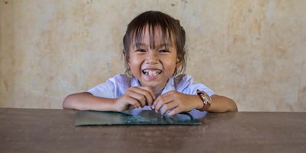 A young girl poses with a big smile on her face while sitting at a table in Cambodia. A young girl poses with a big smile on her face while sitting at a table in Cambodia.