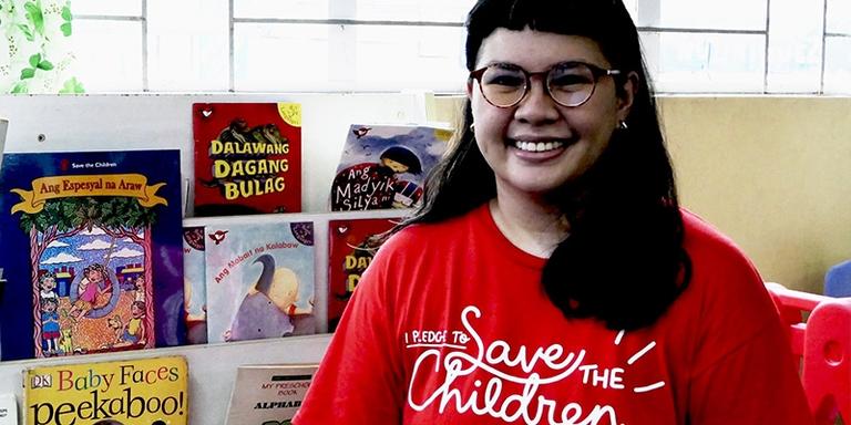 Save the Children worker smiles at the camera in front of a shelf filled with children's books. Save the Children worker smiles at the camera in front of a shelf filled with children's books.