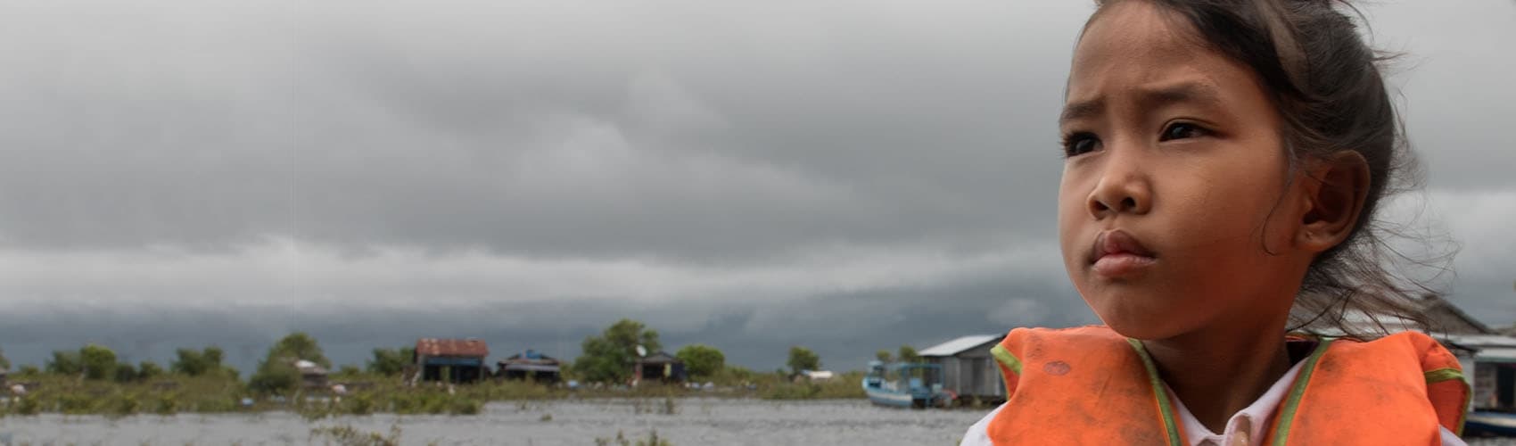 A girl in an orange life jacket stands in front of a small village, set on a river. 