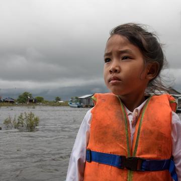 A girl in an orange life jacket stands in front of a village, set on a river. A girl in an orange life jacket stands in front of a village, set on a river.