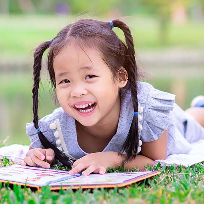 A little girl reads a book in the grass, while smiling at the camera
