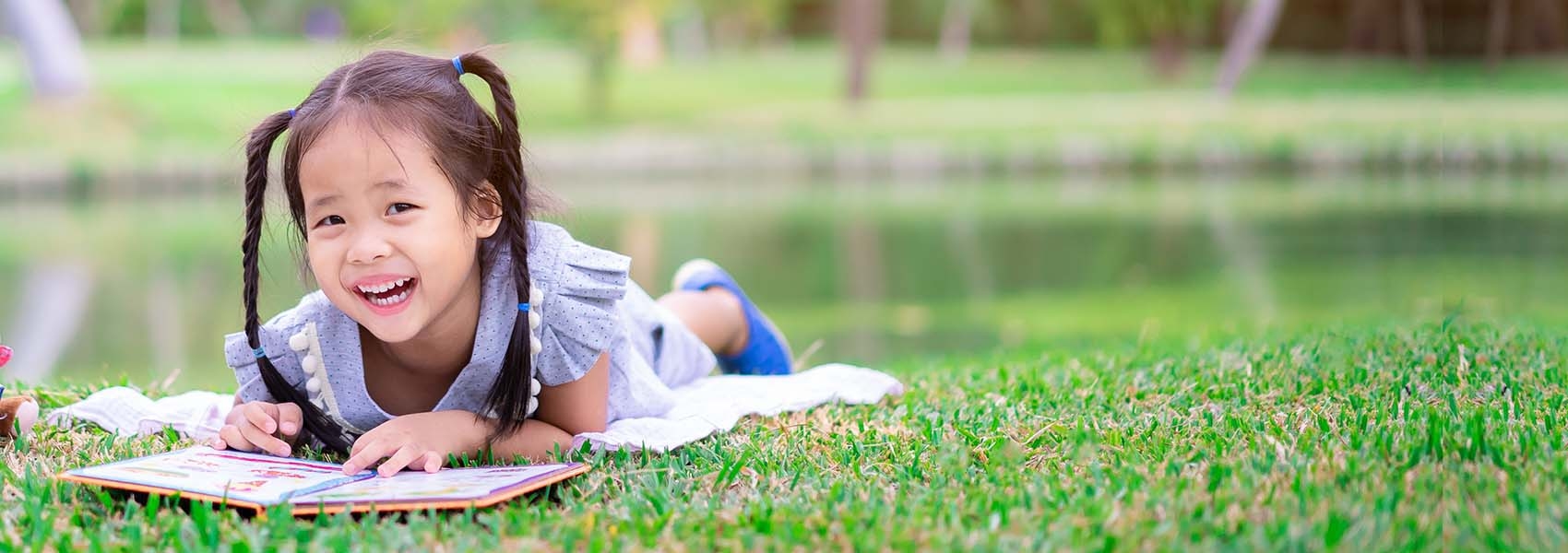 A little girl reads a book in the grass, while smiling at the camera