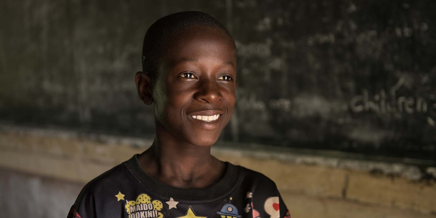 Malawi, a boy in a black t-shirt smiles while in front of a chalk board in a classroom. Malawi, a boy in a black t-shirt smiles while in front of a chalk board in a classroom.