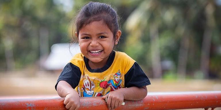 in Indonesia, a young girl smiles while leaning on an outdoor structure. in Indonesia, a young girl smiles while leaning on an outdoor structure.