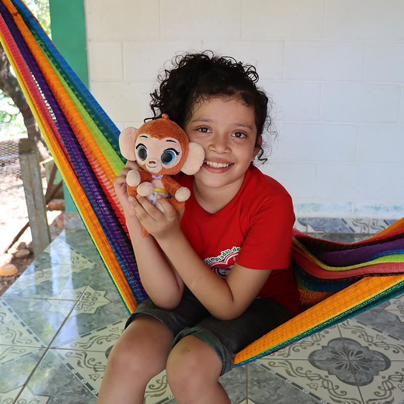 A girl in El Salvador sits in a colorful hammock holding a stuffed monkey