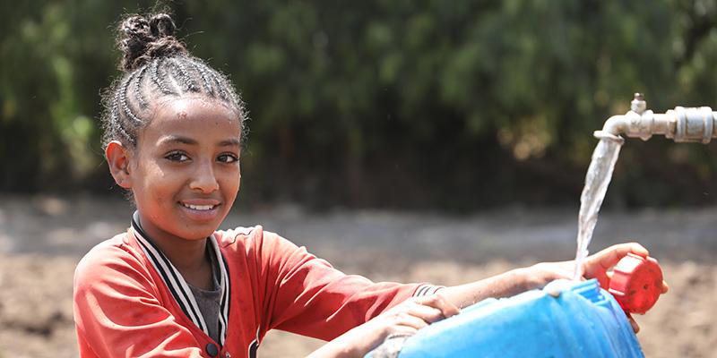 A girl in Ethiopia in an orange shirt fills a jug of water from a tap