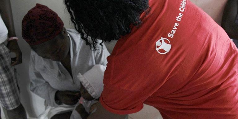 A Save the Children staff member leans over a doctor treating a baby. A Save the Children staff member leans over a doctor treating a baby.