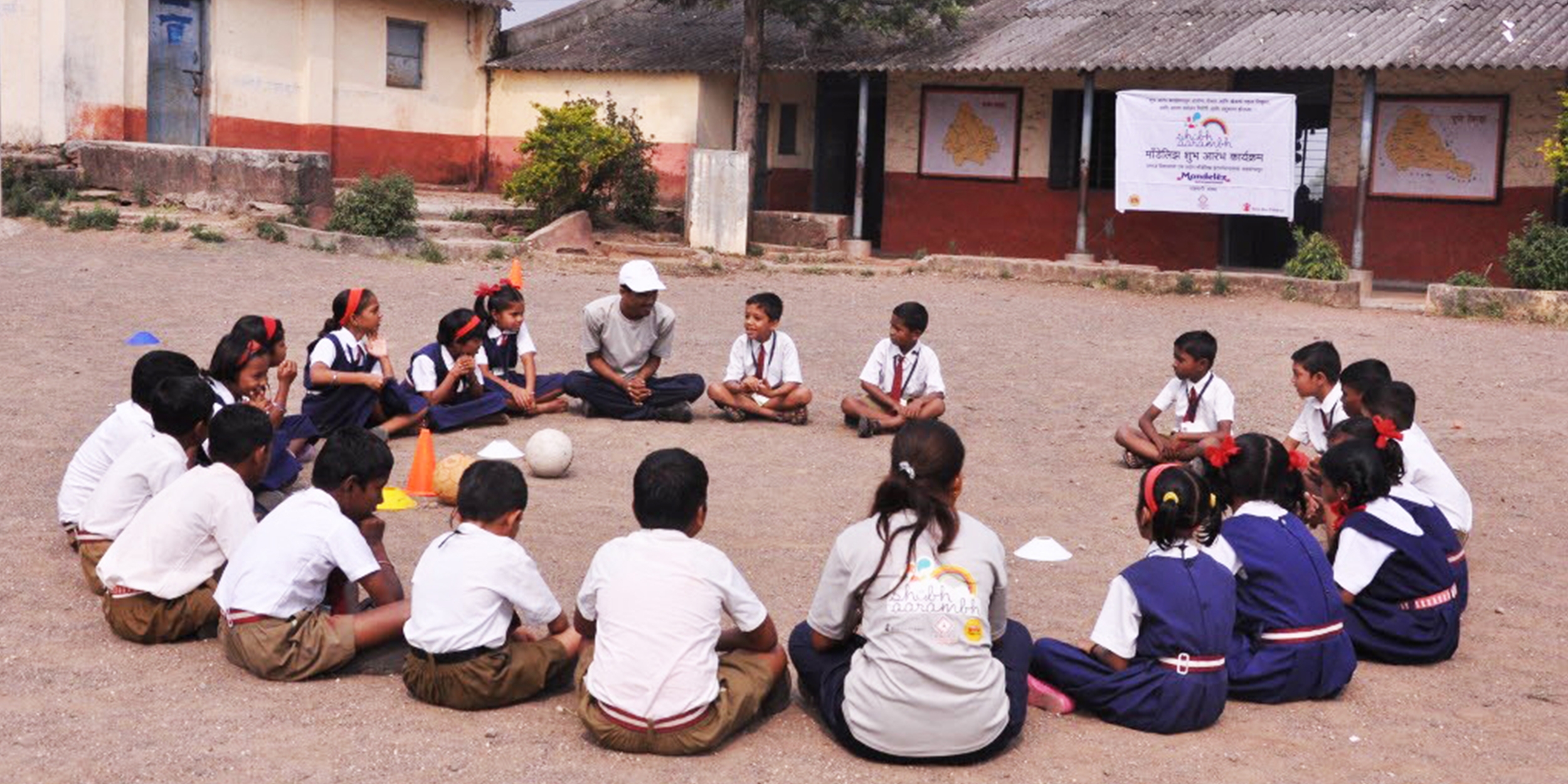 A group of children sit on the ground in a circle at a school in Induri, Maharashtra, India. Their attention is on a teacher. These play sessions are sponsored by Mondelez, and involve learning nutrition and playing games to reinforce a positive message about exercise and good nutrition. Photo credit: Luciana Bonifacio / Save the Children, November 2015.