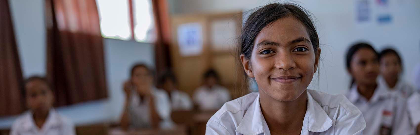 A young girl sits at a desk and smiles at the camera.