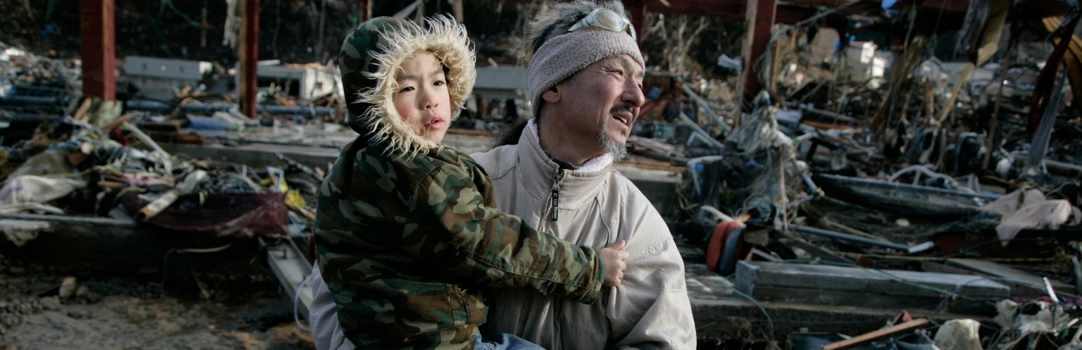 A Japanese man, holding his 6-year-old son, walks through streets filled with debris after two tsunami waves devastated their town of Onigawa, Japan in 2011. They escaped, but their home was hit by a 10-meter high wave; nothing was left of it. Photo credit: Jensen Walker/Save the Children, March 2011.