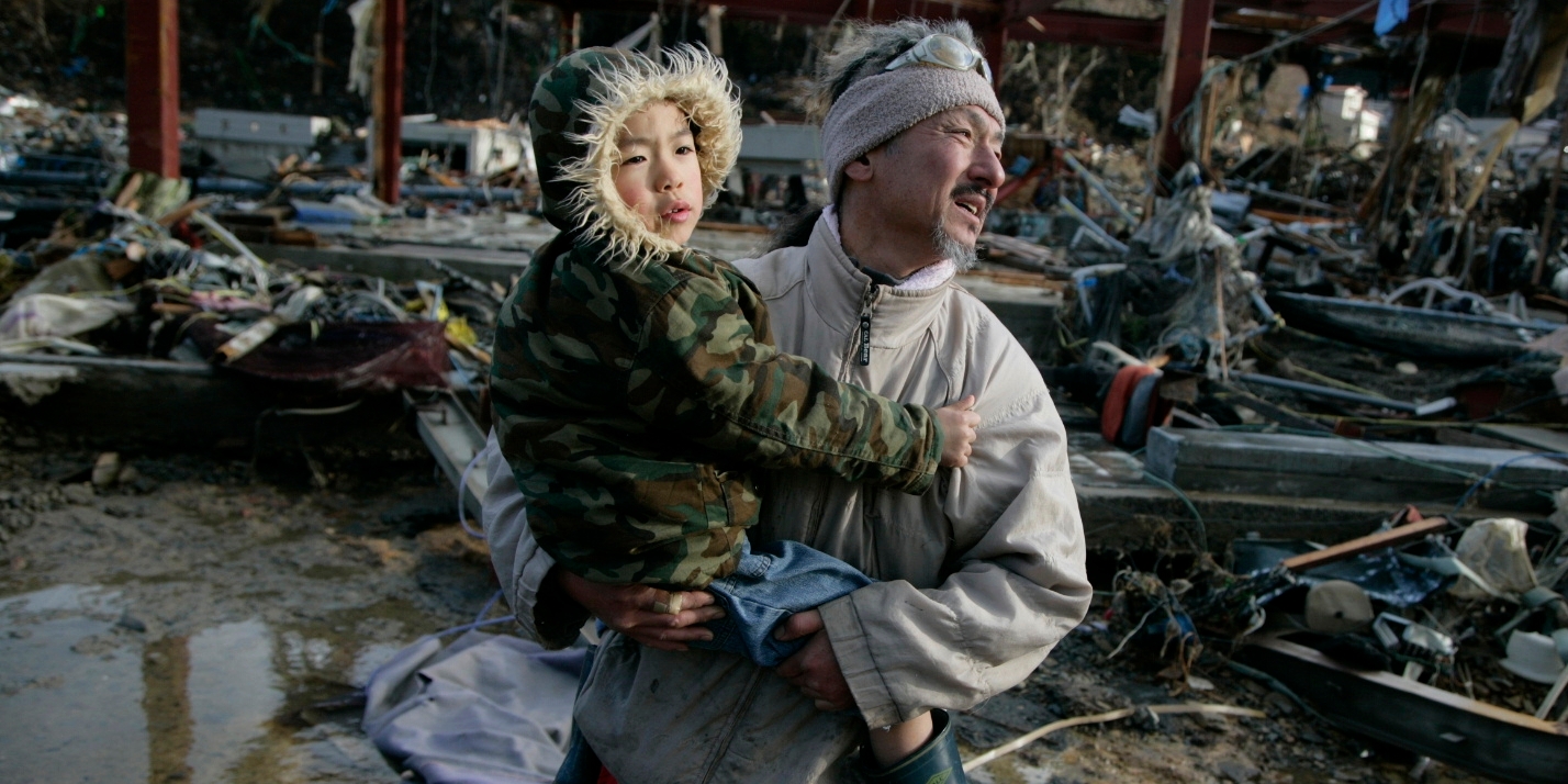 A Japanese man, holding his 6-year-old son, walks through streets filled with debris after two tsunami waves devastated their town of Onigawa, Japan in 2011. They escaped, but their home was hit by a 10-meter high wave; nothing was left of it. Photo credit: Jensen Walker/Save the Children, March 2011.