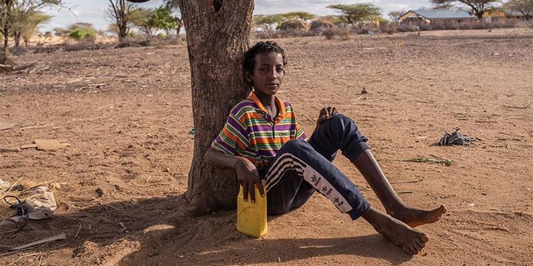 A young boy in Kenya sits against a tree in the desert. A young boy in Kenya sits against a tree in the desert.