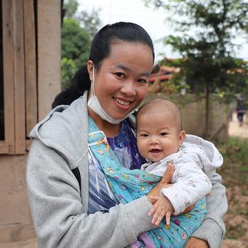 A woman holds her baby and smiles at the camera while receiving emergency food in Laos after a flood. A woman holds her baby and smiles at the camera while receiving emergency food in Laos after a flood.