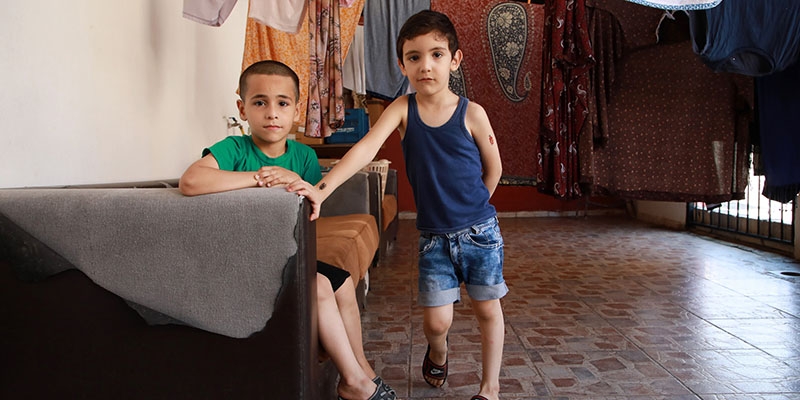 Two boys pose in an empty room. One sits on a couch, the other leans against it. Two boys pose in an empty room. One sits on a couch, the other leans against it.