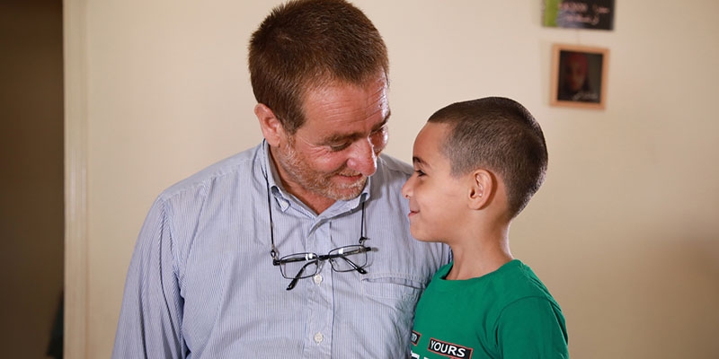Farid and his son stand next to and look toward each other, smiling. Farid and his son stand next to and look toward each other, smiling.