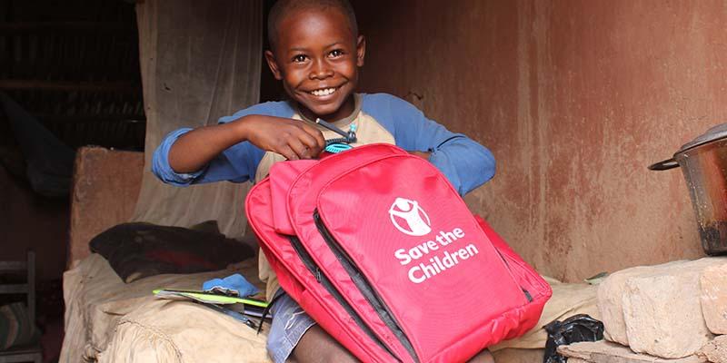 In Madagascar, a boy holds a red backpack with a Save the Children logo on it. 