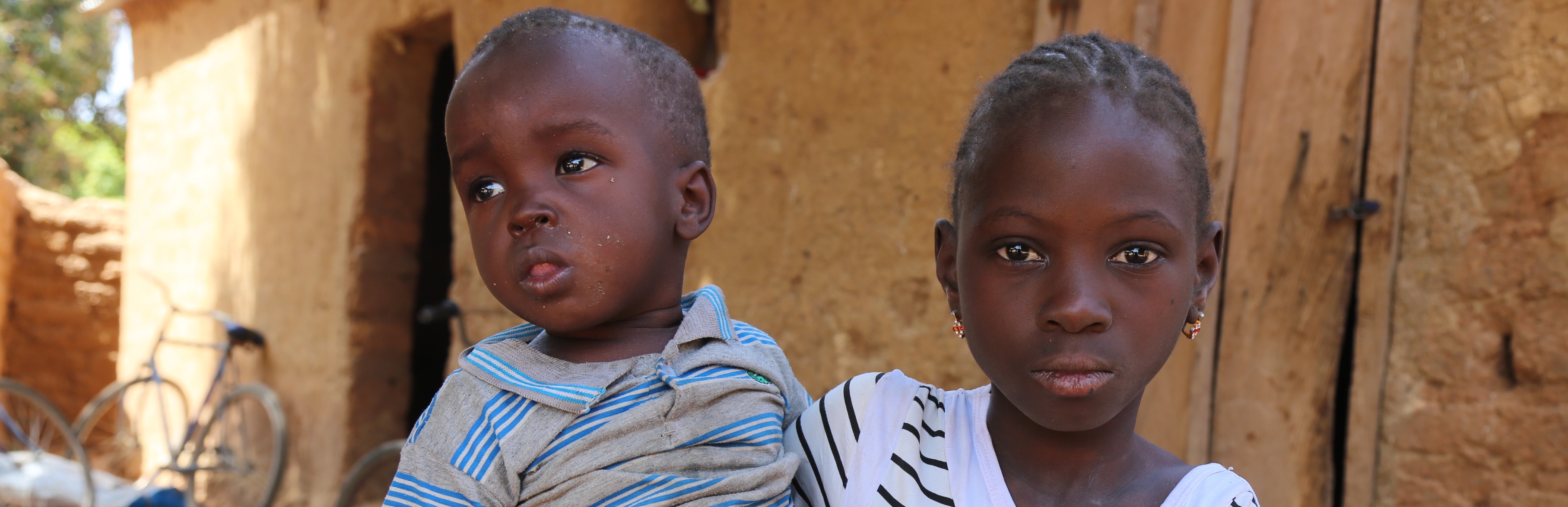 An 8-year old girl holds her baby brother while sitting outside her family's home in Mali.