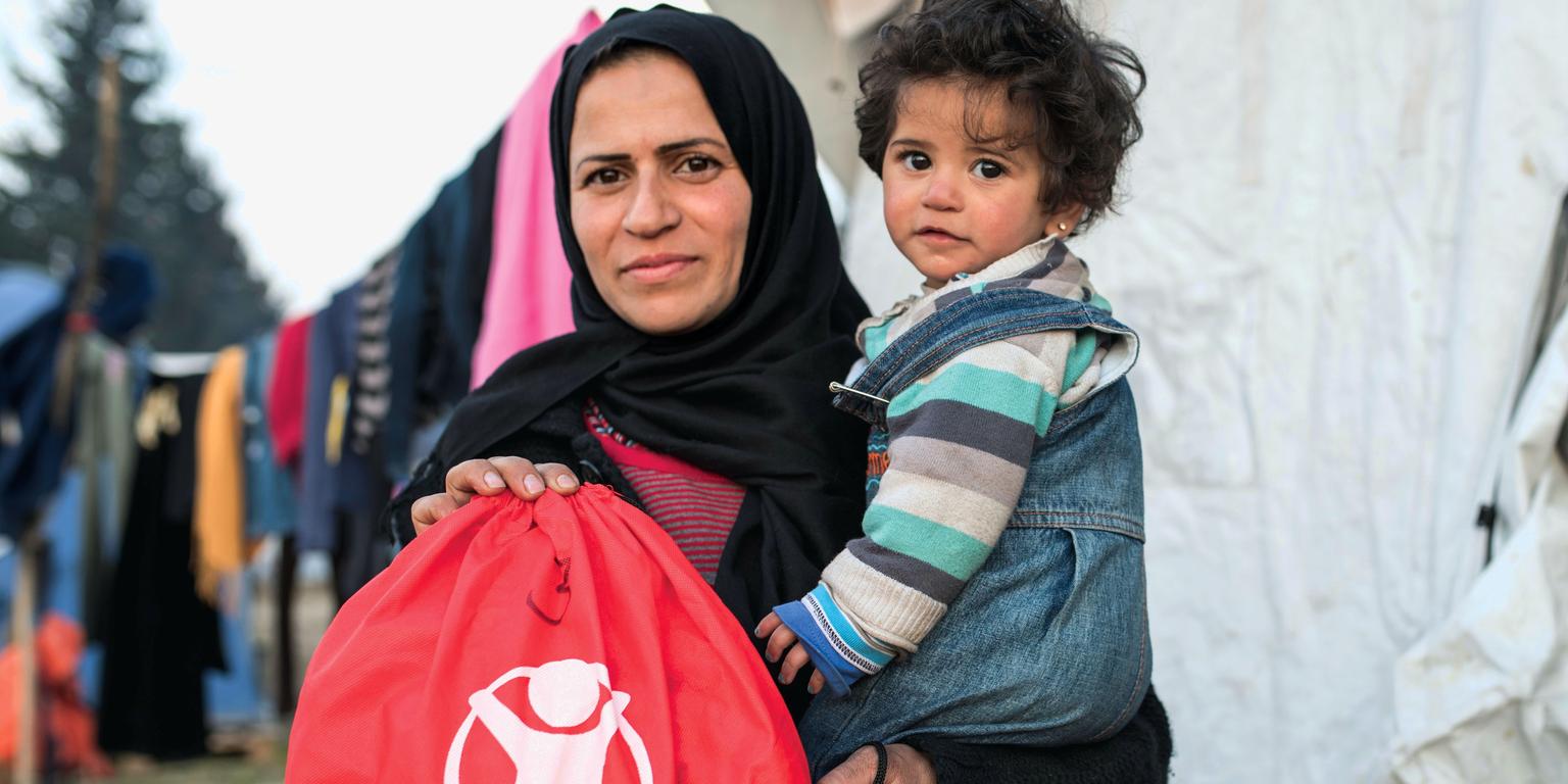 A mother in the Middle East holds her child and a red refugee basics bag from Save the Children relief efforts A mother in the Middle East holds her child and a red refugee basics bag from Save the Children relief efforts