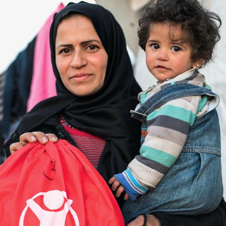 A caregiver holds a baby and a Save the Children backpack. A caregiver holds a baby and a Save the Children backpack.