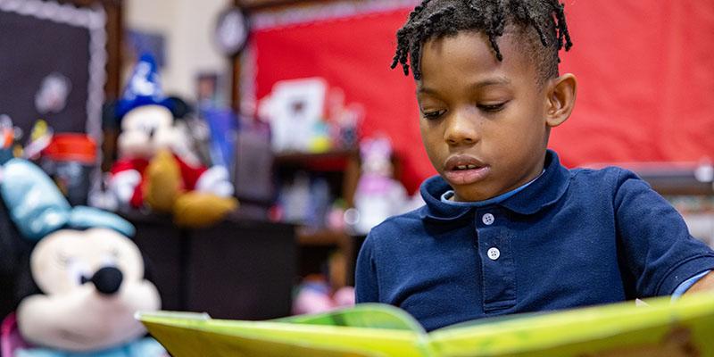 An 8-year-old boy in a navy-blue shirt reads with in a Save the Children room. 