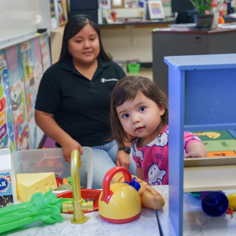 Little girl, 1, prepares lunch in a play kitchen with Lead Associate of Early Childhood Programs