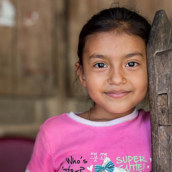 Six-year-old Manela, wearing a pink shirt, smiles as she leans against a wooden door. Six-year-old Manela, wearing a pink shirt, smiles as she leans against a wooden door.