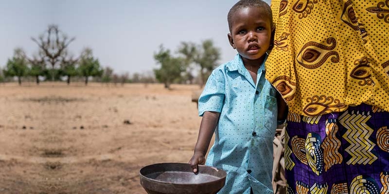 In Nigeria, a young boy hugs his mother's leg while standing outside and holding a bucket. In Nigeria, a young boy hugs his mother's leg while standing outside and holding a bucket.