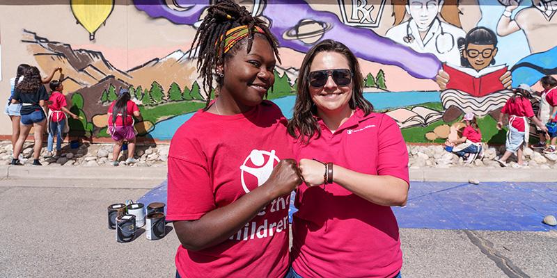 NM, Save the Children staff members, fist bump while standing in front of a colorful mural.
