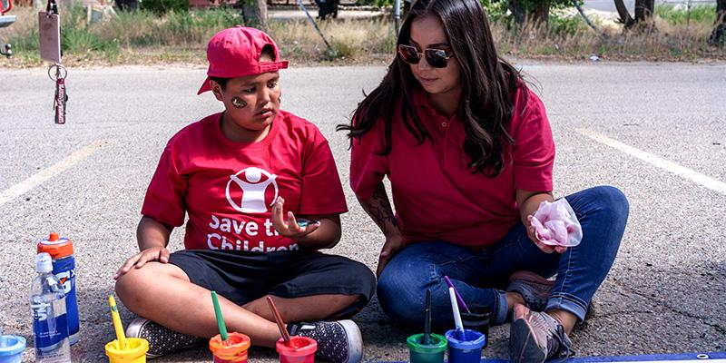 NM, a Save the Children staff member sits on the ground painting with a child. -ch1849978-rec
