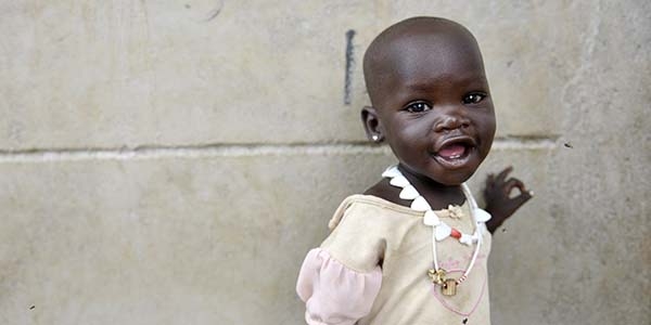 A one-year-old smiles as she places one hand on a wall near a food distribution center in South Sudan. A one-year-old smiles as she places one hand on a wall near a food distribution center in South Sudan.
