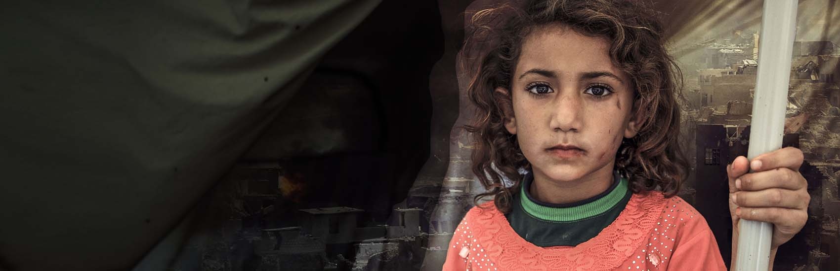 A Syrian girl stands in front of a makeshift shelter as a scene of fire and destruction is shown in the background.