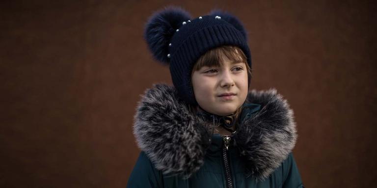 In Ukraine, a girl stands outside alone on a dreay day while wearing a dark coat and hat. In Ukraine, a girl stands outside alone on a dreay day while wearing a dark coat and hat.