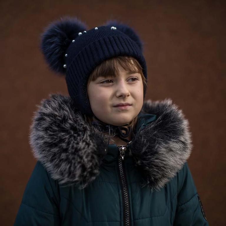 In Ukraine, a girl dressed in a hat and coat stands alone against a dark background. In Ukraine, a girl dressed in a hat and coat stands alone against a dark background.