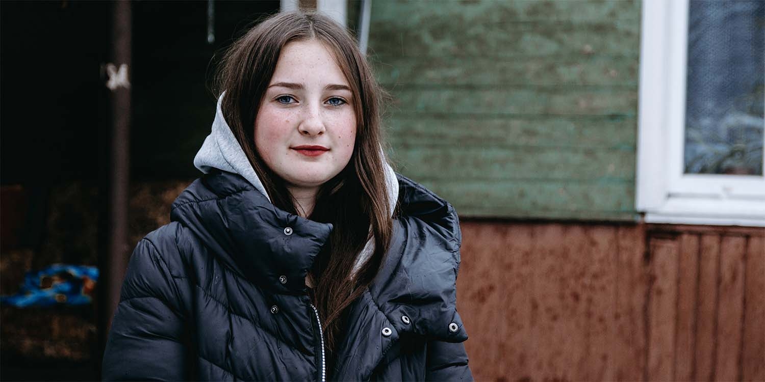 In Ukraine, a girl dressed in a hat and coat stands alone against a dark background. In Ukraine, a girl dressed in a hat and coat stands alone against a dark background.