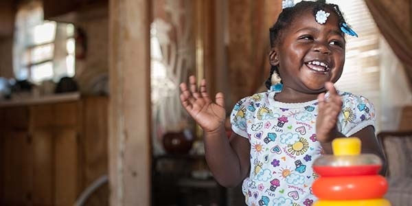 A 20-month old girl who lives in Mississippi smiles as she gets ready to clap her hands while standing near a table with a bright toy on it. A 20-month old girl who lives in Mississippi smiles as she gets ready to clap her hands while standing near a table with a bright toy on it.