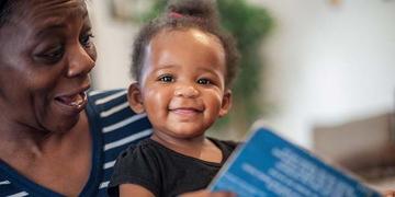 A young child smiles as she holds a picture book while sitting on the lap of an older adult caretaker in her family. A young child smiles as she holds a picture book while sitting on the lap of an older adult caretaker in her family.