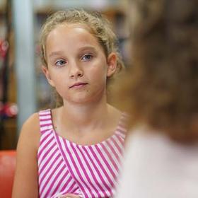 A young girl sits at a table in a library and listens intently to an adult across the table. A young girl sits at a table in a library and listens intently to an adult across the table.