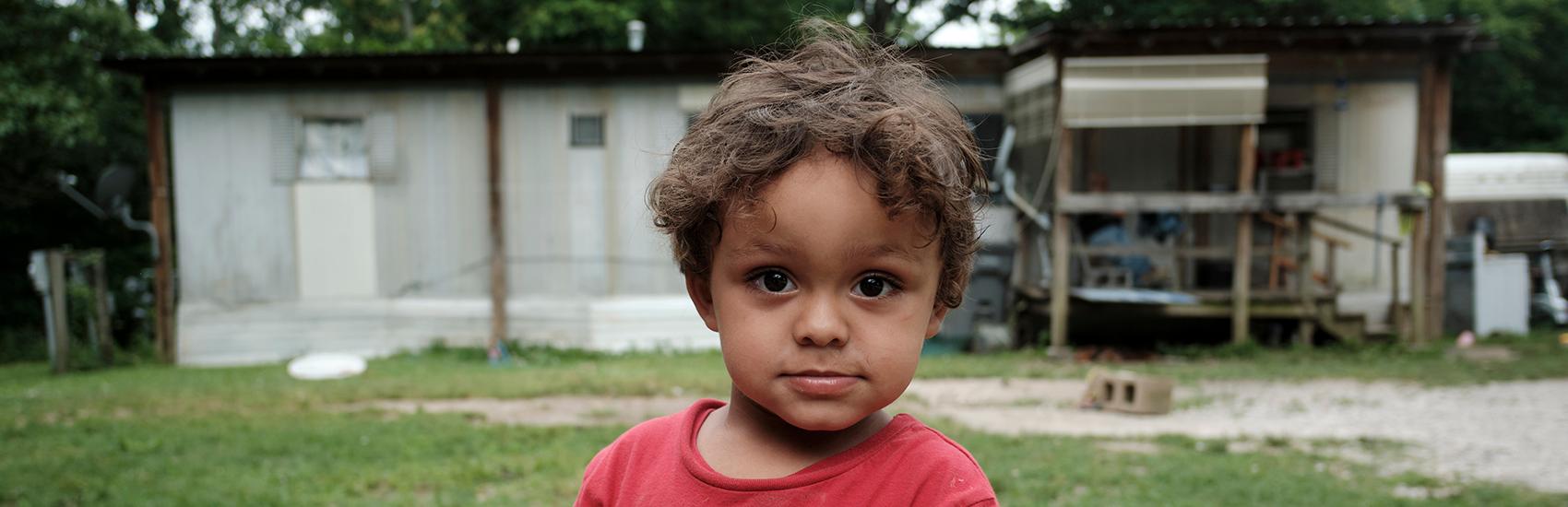 A young child stands in front of their home during a home visit from Save the Children to deliver food.