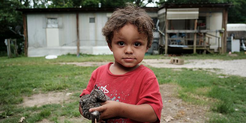 A young child stands in front of their home during a home visit from Save the Children to deliver food. A young child stands in front of their home during a home visit from Save the Children to deliver food.