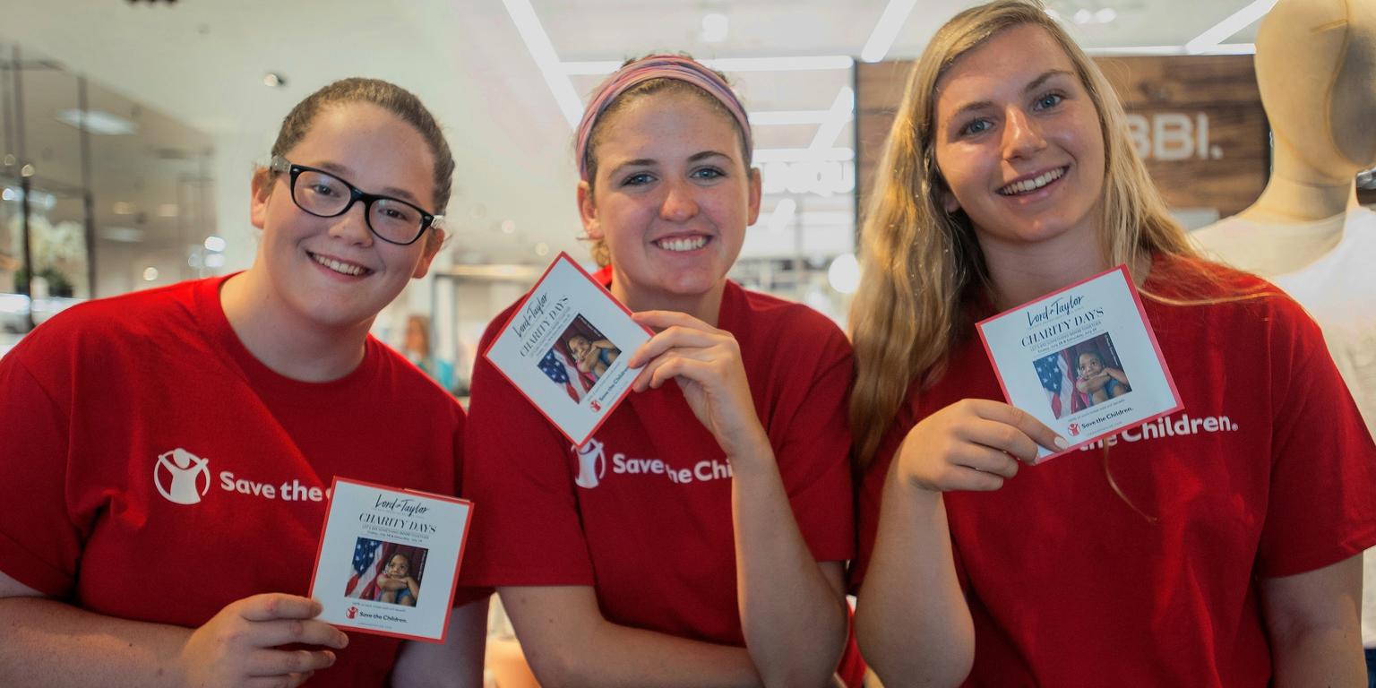 Three women wearing red Save the Children shirts and holding campaign cards gather together at a Save the Children store event. The women are volunteers helping to promote a campaign throughout the store. Photo credit: Susan Warner/Save the Children, July 2017. Three women wearing red Save the Children shirts and holding campaign cards gather together at a Save the Children store event. The women are volunteers helping to promote a campaign throughout the store. Photo credit: Susan Warner/Save the Children, July 2017.