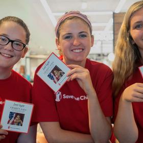Three women wearing red Save the Children shirts and holding campaign cards gather together at a Save the Children store event. The women are volunteers helping to promote a campaign throughout the store. Photo credit: Susan Warner/Save the Children, July 2017. Three women wearing red Save the Children shirts and holding campaign cards gather together at a Save the Children store event. The women are volunteers helping to promote a campaign throughout the store. Photo credit: Susan Warner/Save the Children, July 2017.