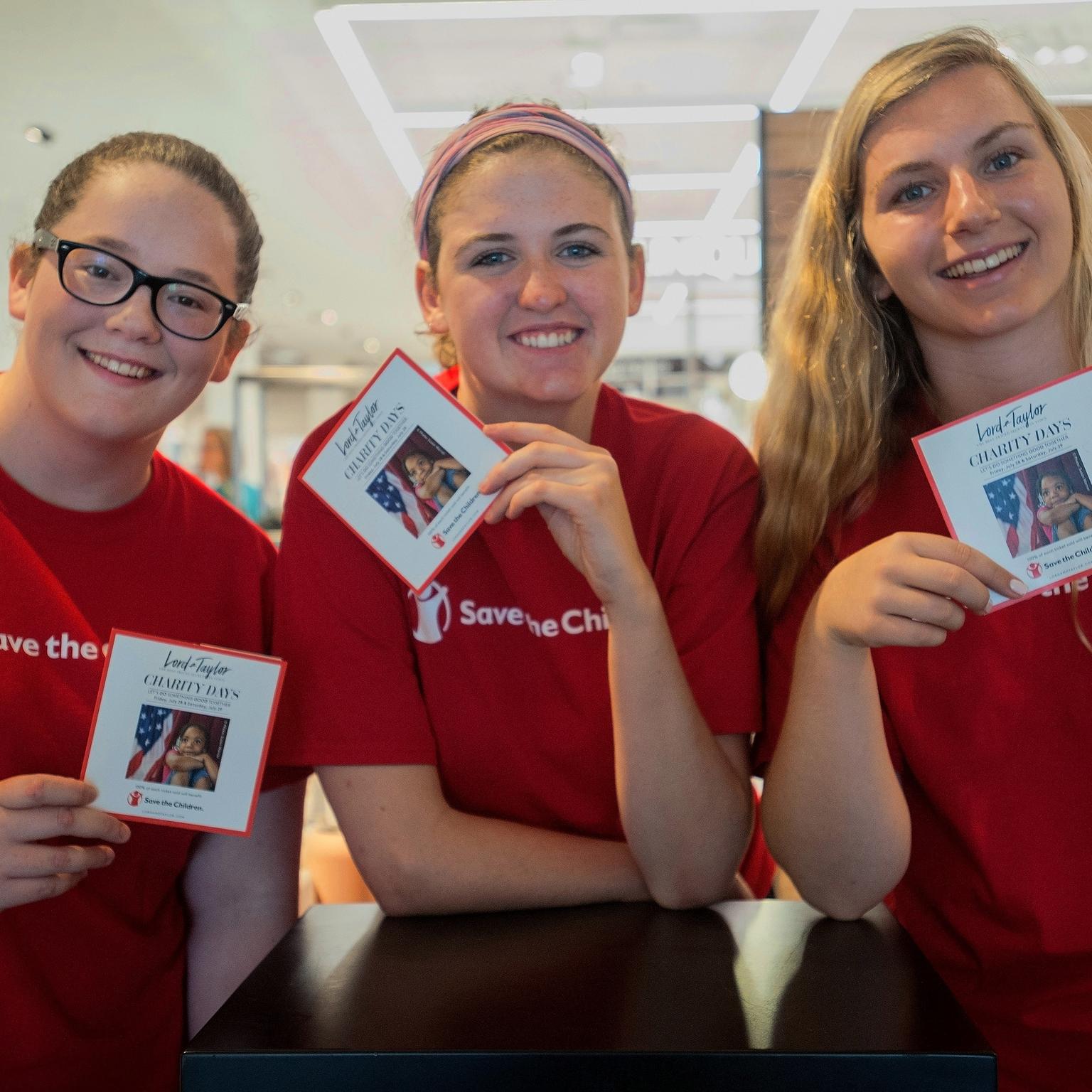 Three women wearing red Save the Children shirts and holding campaign cards gather together at a Save the Children store event. The women are volunteers helping to promote a campaign throughout the store. Photo credit: Susan Warner/Save the Children, July 2017. Three women wearing red Save the Children shirts and holding campaign cards gather together at a Save the Children store event. The women are volunteers helping to promote a campaign throughout the store. Photo credit: Susan Warner/Save the Children, July 2017.